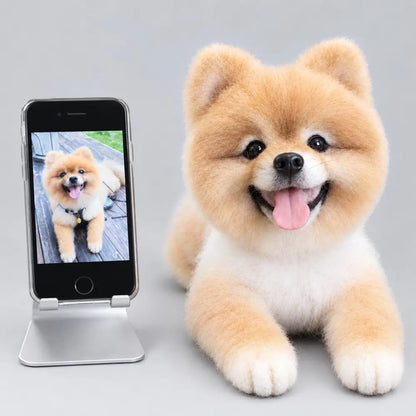 Custom felted tan Pomeranian sculpture lying with its front paws forward and tongue out, beside the reference dog photo on a phone.