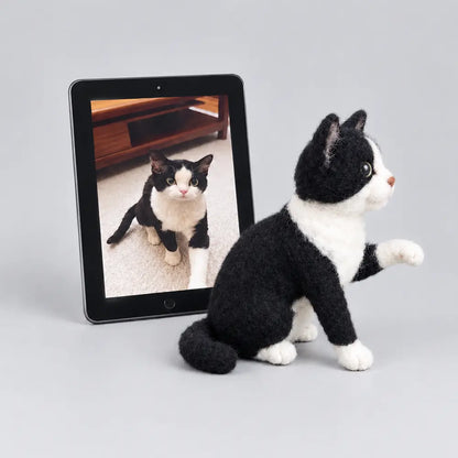 Custom felted tuxedo cat sculpture with one front paw raised, shown in side view beside the original cat photo on a tablet.
