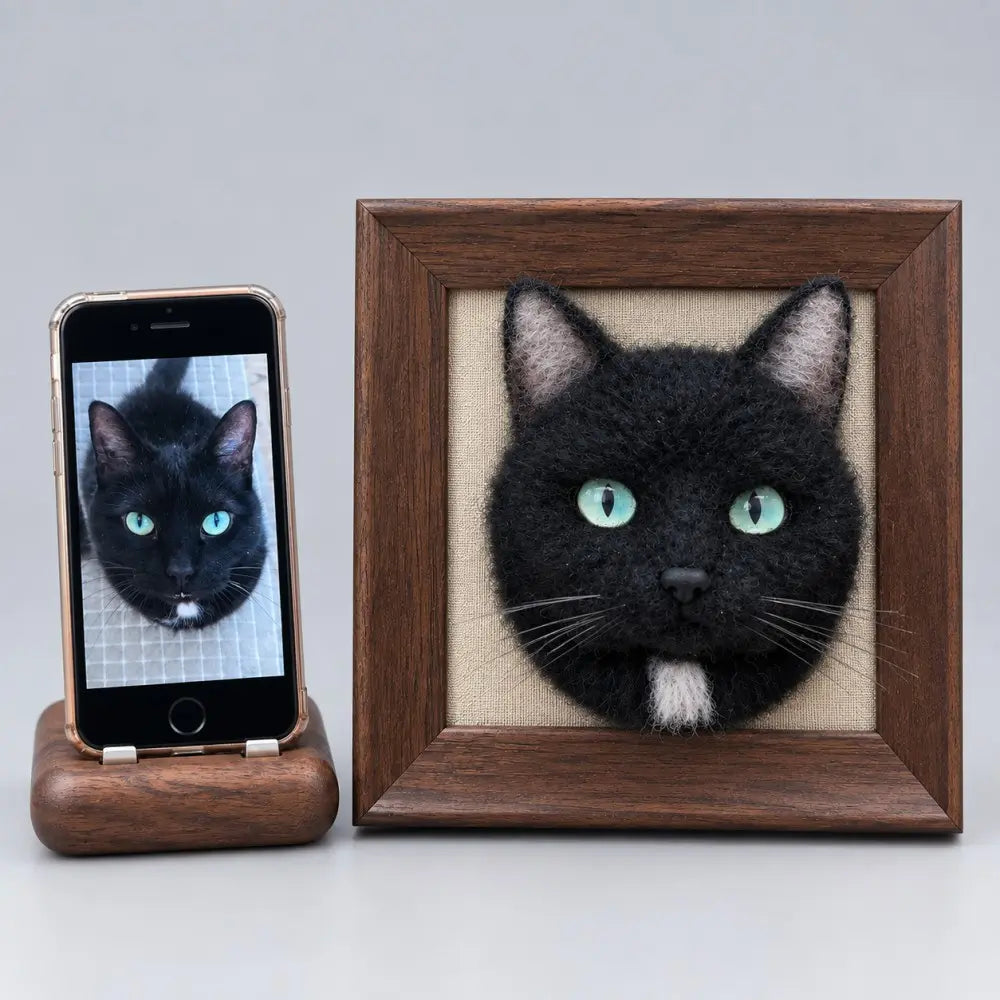 Custom 3D needle felted black cat portrait with green eyes and white chin in a square wooden frame next to a smartphone showing the original cat photo